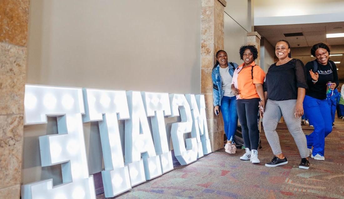 Four M州 students laugh 和 pose in front of a large light-up M州 sign